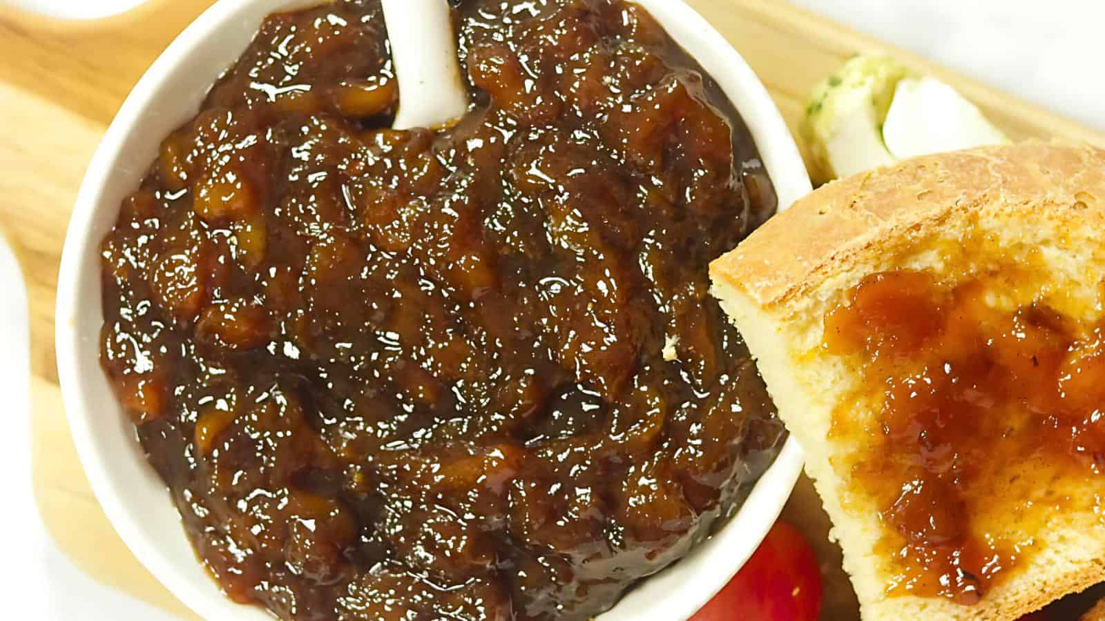 A small white bowl filled with dark brown onion marmalade, featuring a spoon inside. Beside the bowl is a slice of toast generously topped with the same marmalade. The background shows more toast and a hint of green garnish.