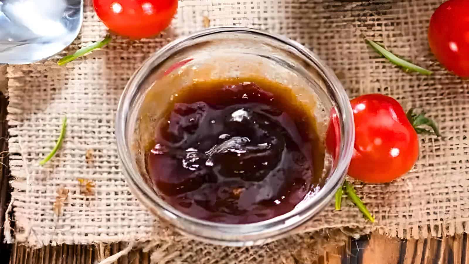 A glass jar containing a dark, thick sauce is placed on a burlap surface with cherry tomatoes and rosemary sprigs nearby.