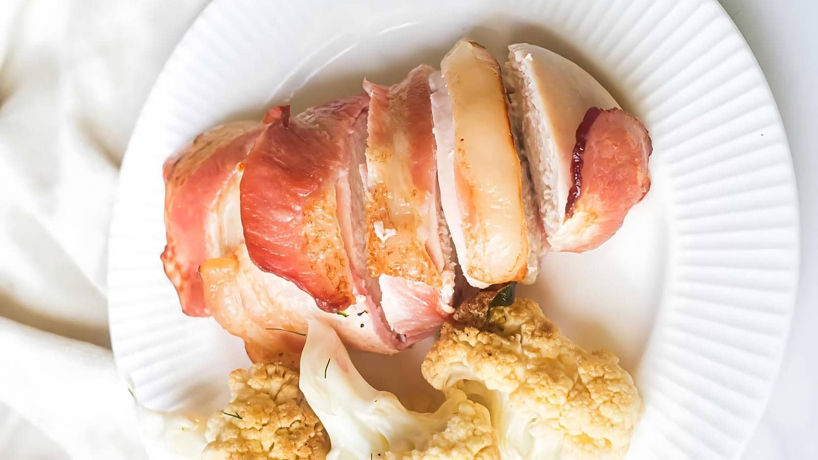 A plate with sliced bacon-wrapped chicken breast and roasted cauliflower florets.