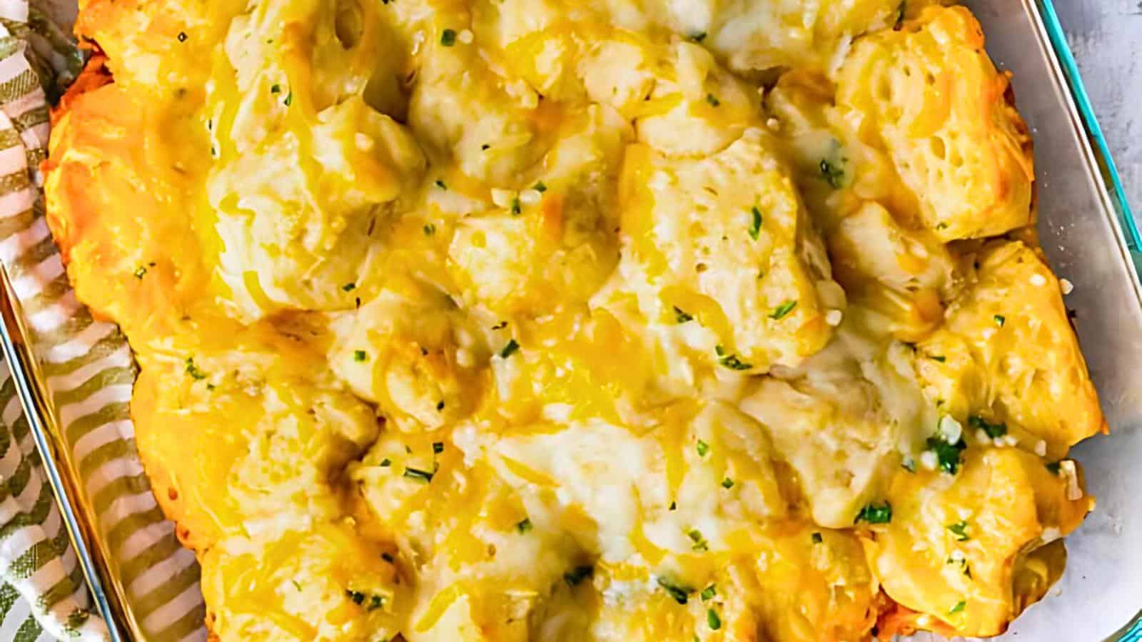 A close-up of a casserole dish filled with golden, baked biscuit pieces topped with melted cheese and garnished with chopped herbs. The dish is placed next to a striped cloth on a tabletop.