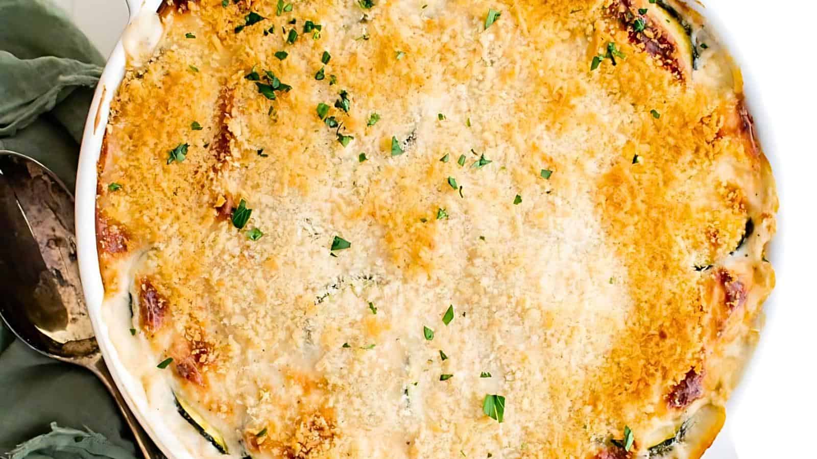 A baked casserole topped with a golden-brown breadcrumb crust, garnished with chopped parsley. A serving spoon and green cloth are partially visible on the left side.