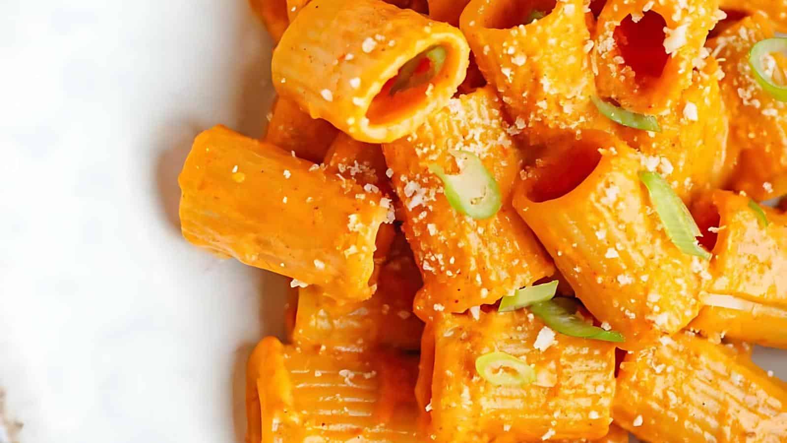 Close-up of rigatoni pasta coated in a rich, creamy orange sauce, garnished with finely chopped green onions and sprinkled with grated cheese. The dish is presented on a white plate, showcasing the texture and vibrant color of the sauce.