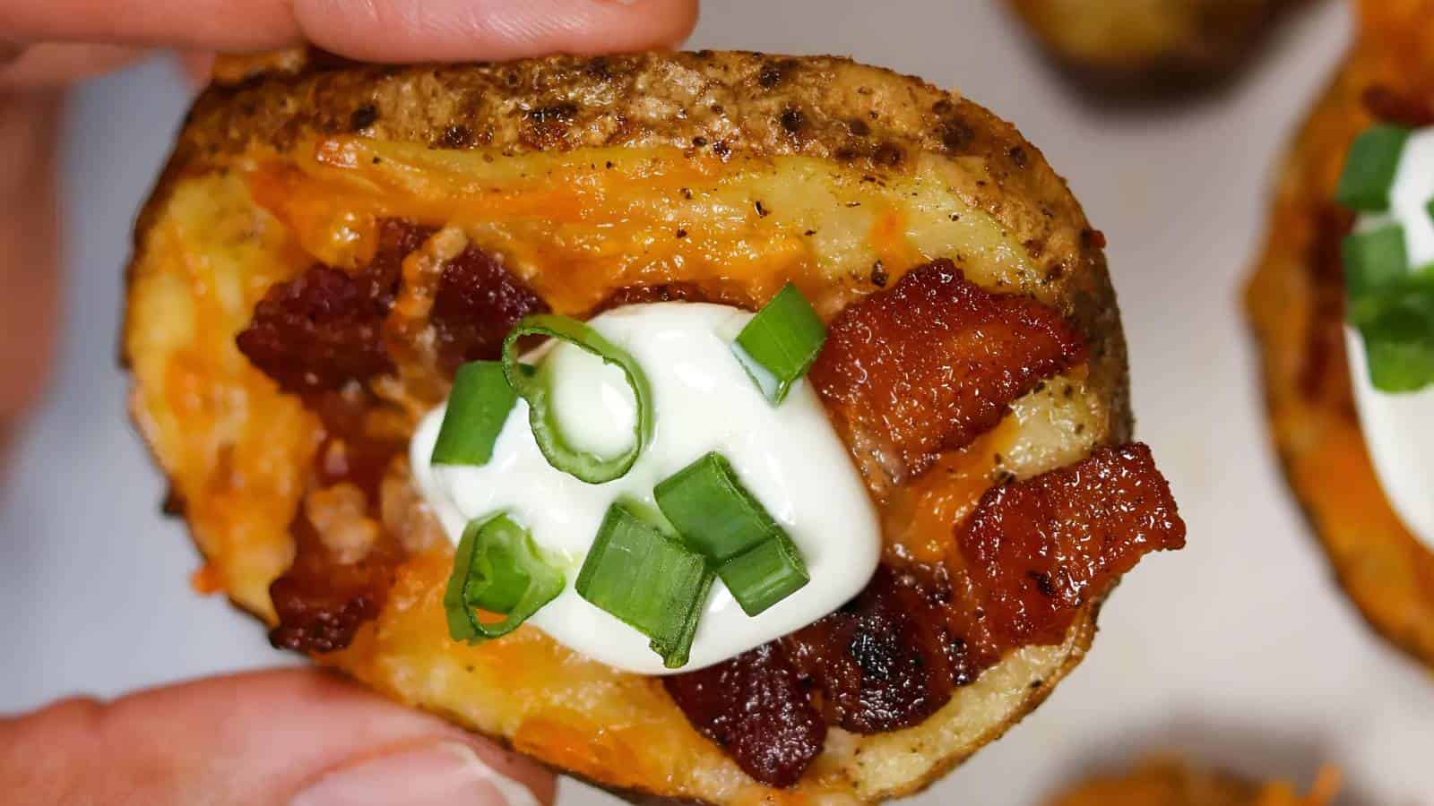 A hand holding a potato skin with melted cheese, bacon pieces, a dollop of sour cream, and chopped green onions.
