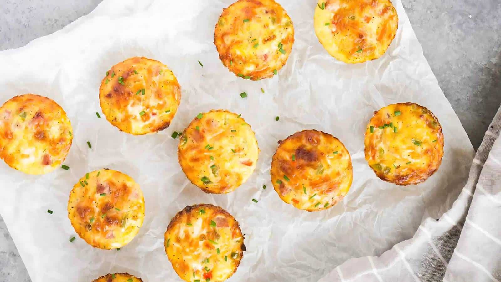 Mini egg muffins with melted cheese and chives are arranged on crumpled parchment paper. The muffins have a golden-brown top and are positioned on a light gray surface. A striped cloth napkin is partially visible in the bottom right corner.