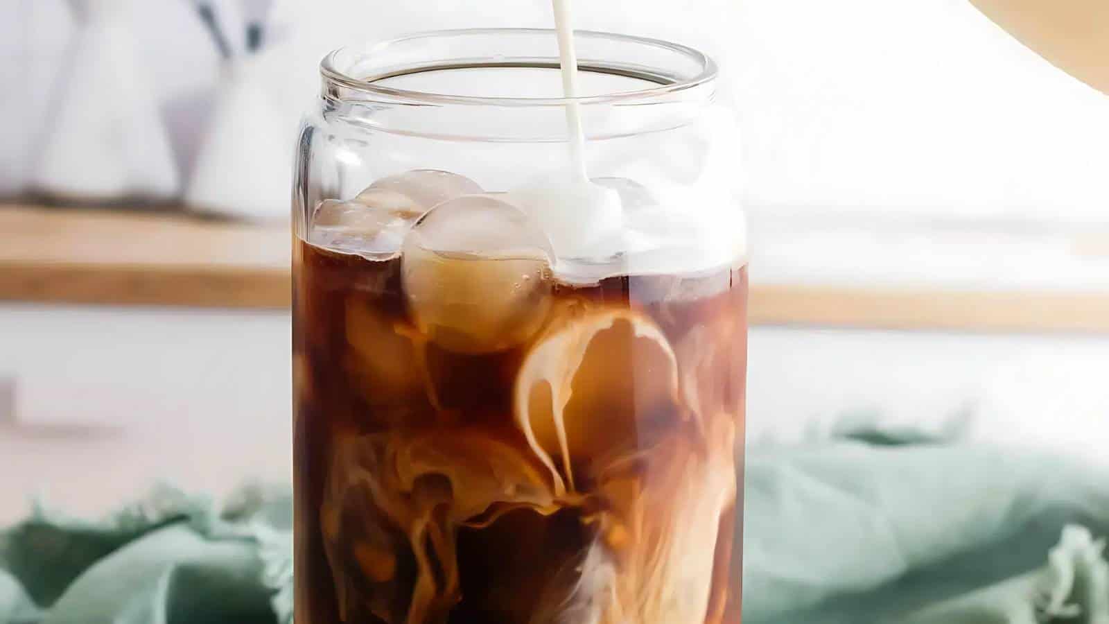 A glass of iced coffee with cream swirling in, on a light green cloth.