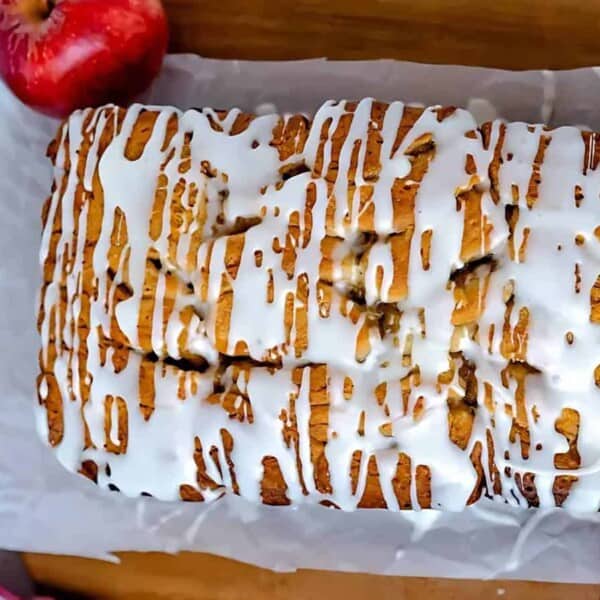 A loaf cake drizzled with white icing sits on a parchment-lined wooden cutting board, surrounded by red apples.