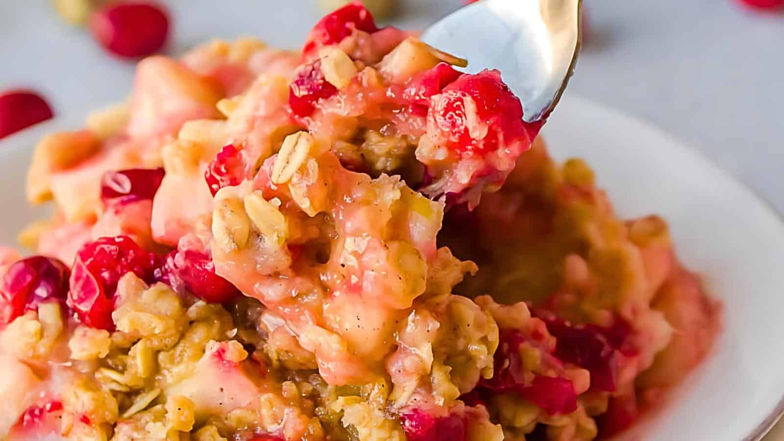 A close-up of a spoonful of baked oatmeal topped with vibrant red cranberries. The oatmeal appears creamy with a mix of textures from oats and fruit, and it is placed in a white bowl with more cranberries scattered around.