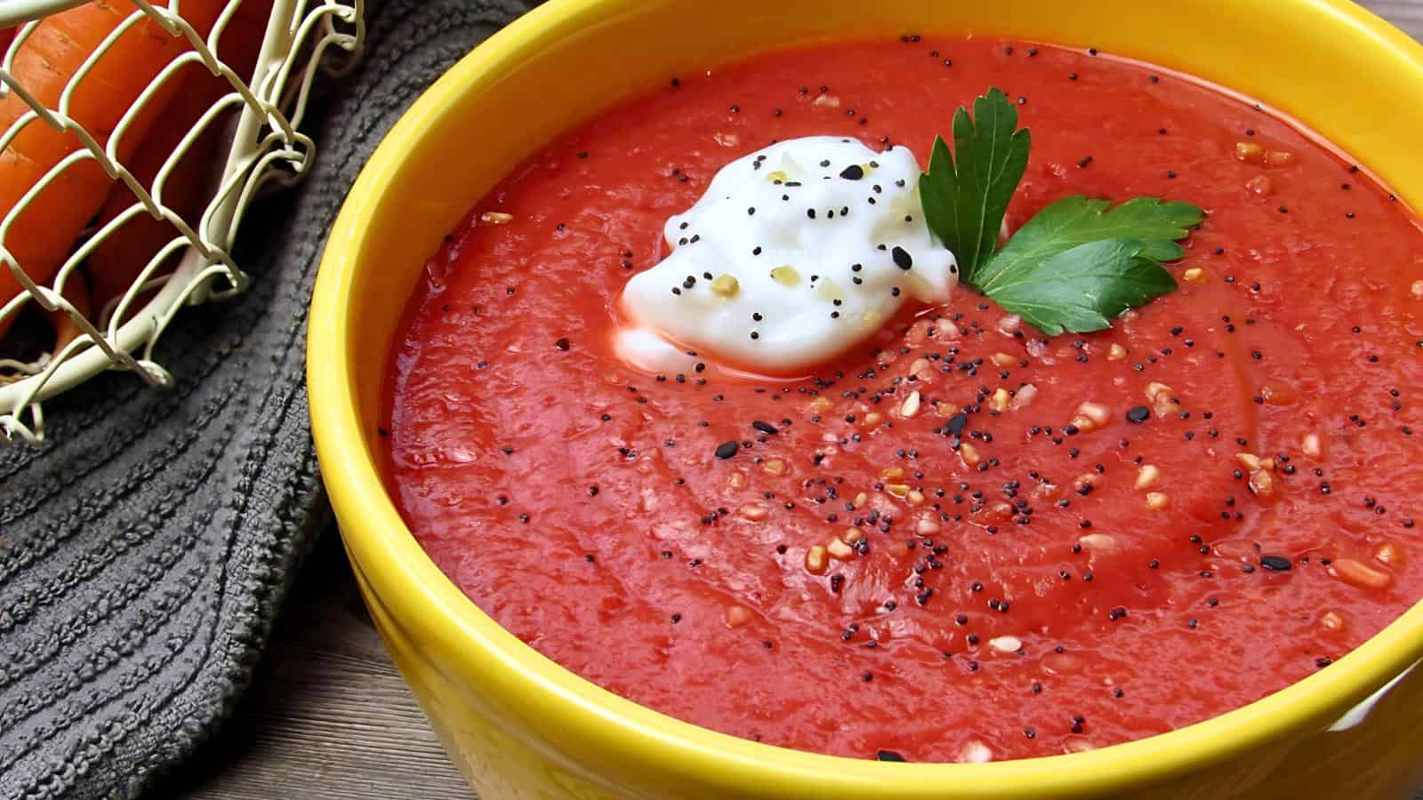 A yellow bowl of tomato soup topped with a dollop of cream and garnished with black sesame seeds and a parsley leaf. A gray cloth and a basket of tomatoes are in the background.