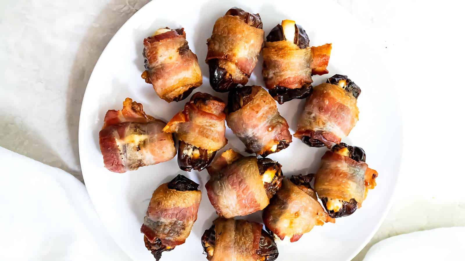 A white plate with several bacon-wrapped dates, each stuffed with cheese. The bacon is crispy and golden brown, and the dates appear glossy and caramelized.