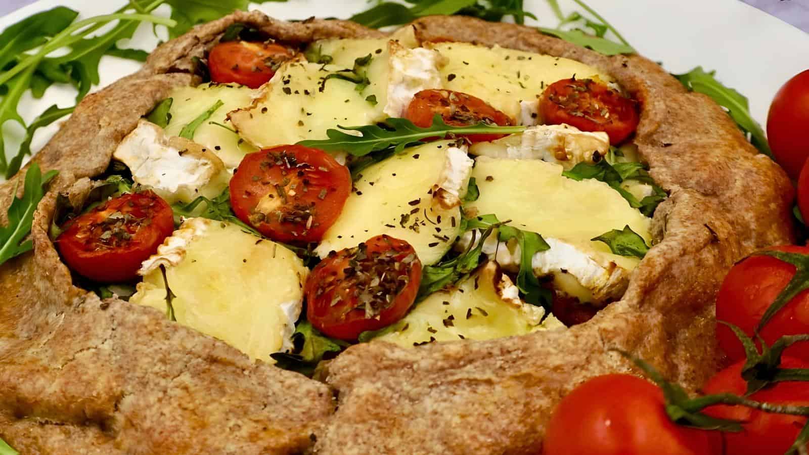 A rustic galette with a whole wheat crust, topped with sliced tomatoes, zucchini, arugula, and herbs. Fresh tomatoes and arugula are placed around the dish.