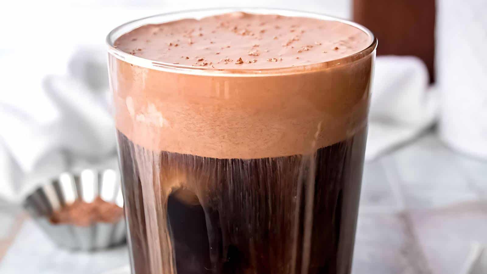 A glass of iced chocolate drink with a frothy top.