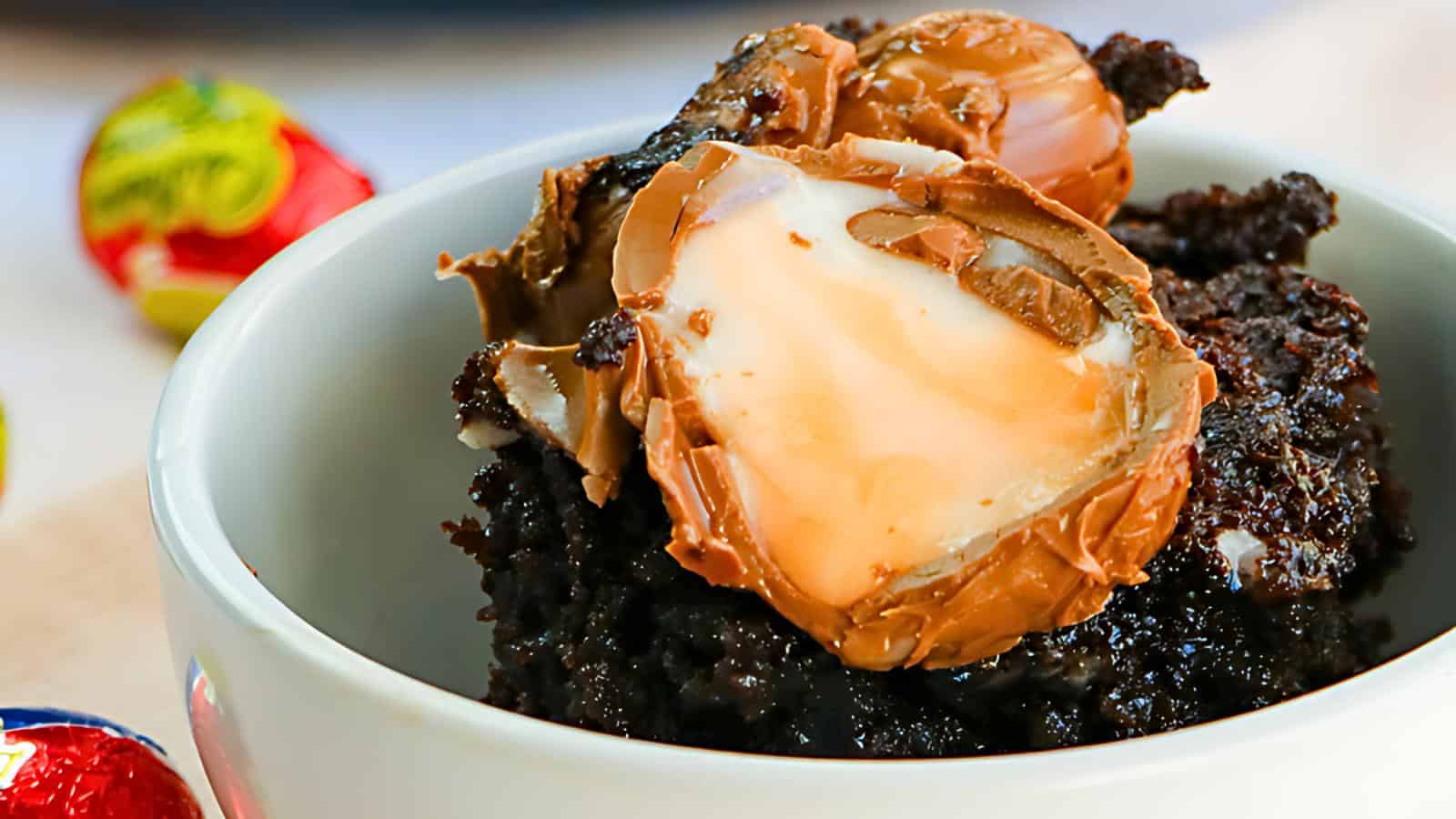 A white bowl containing a piece of chocolate cake topped with a halved chocolate creme egg, surrounded by colorful foil-wrapped eggs.