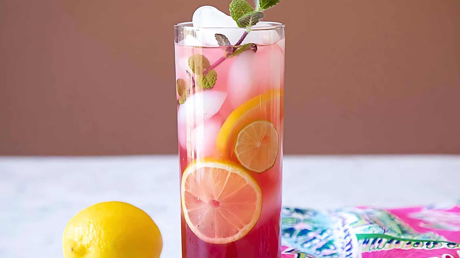 A tall glass with a layered drink, garnished with lemon slices and mint leaves, next to a whole lemon and a pink patterned cloth.