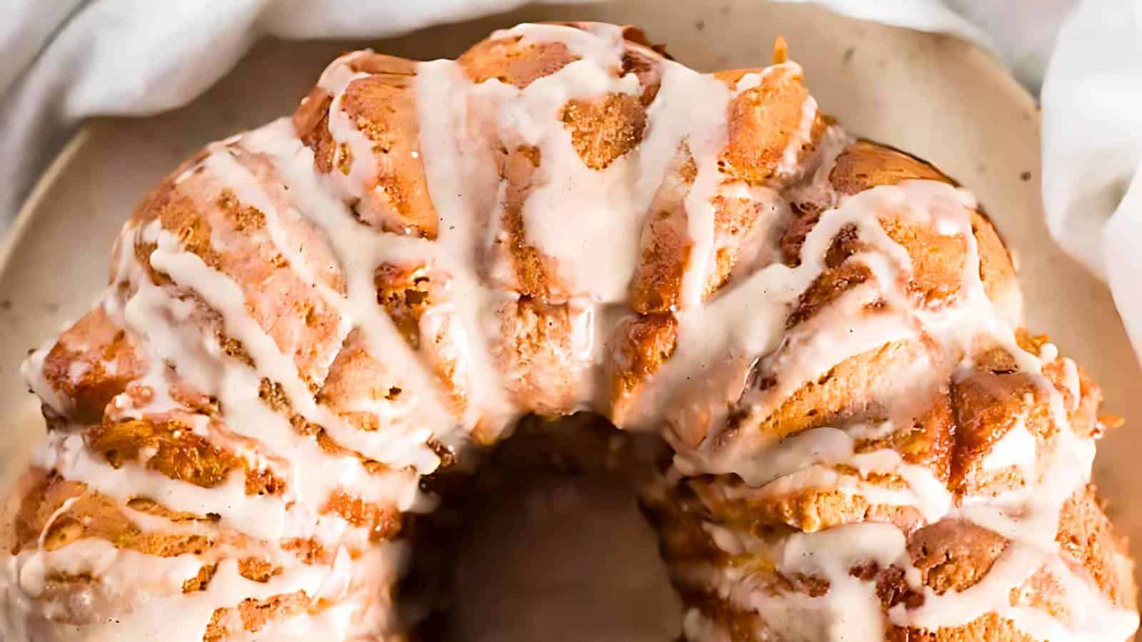 A close-up of a glazed monkey bread, showcasing its golden-brown, pull-apart pieces with drizzled white icing. The bread is on a round plate with a light fabric partially visible in the background.