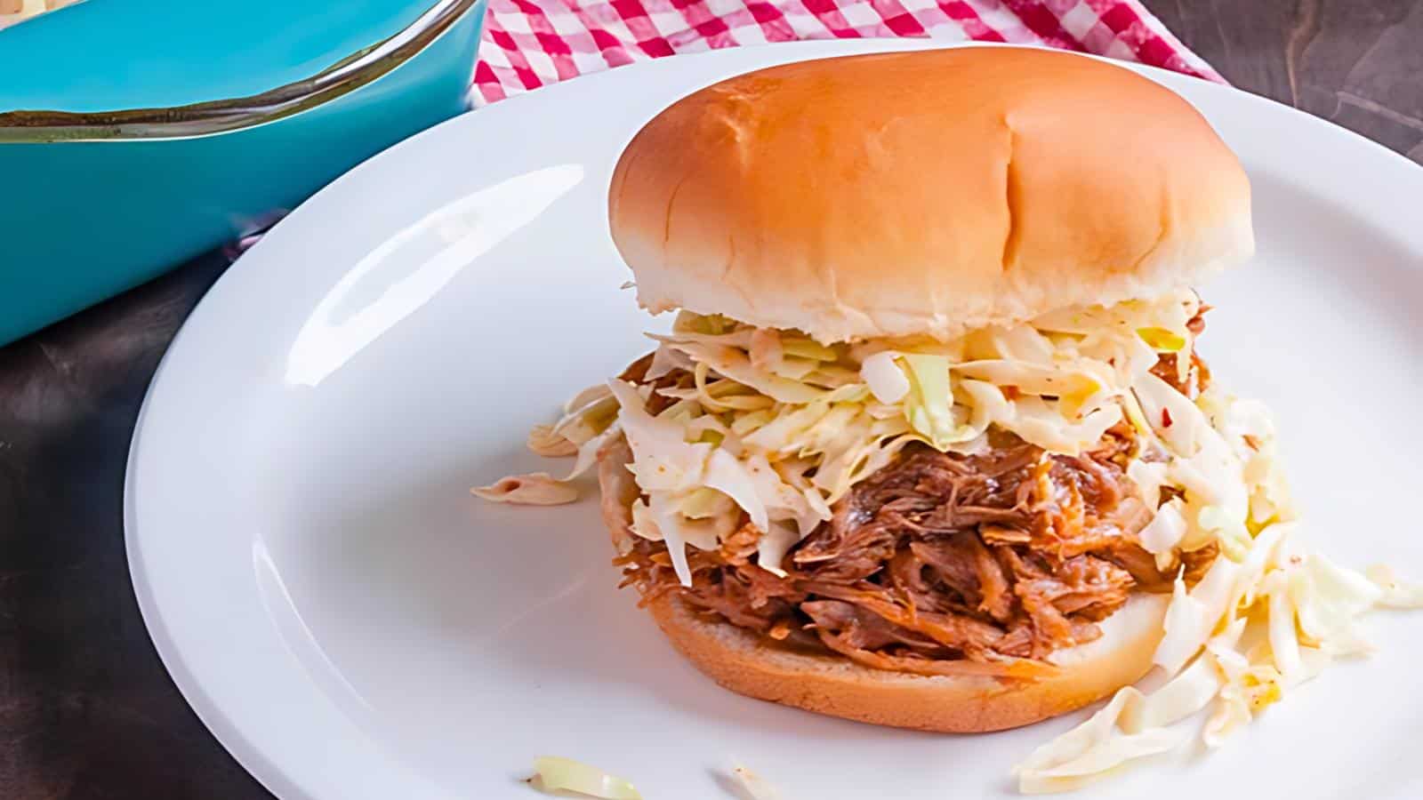 A white plate with a pulled pork sandwich topped with coleslaw, accompanied by a red-checked cloth and a blue dish.