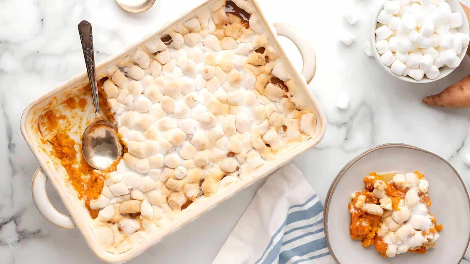 An overhead view of a white baking dish filled with sweet potato casserole topped with toasted marshmallows. A spoon rests inside the dish. Next to it is a plate with a serving of the casserole, a small bowl of marshmallows, and a striped cloth.