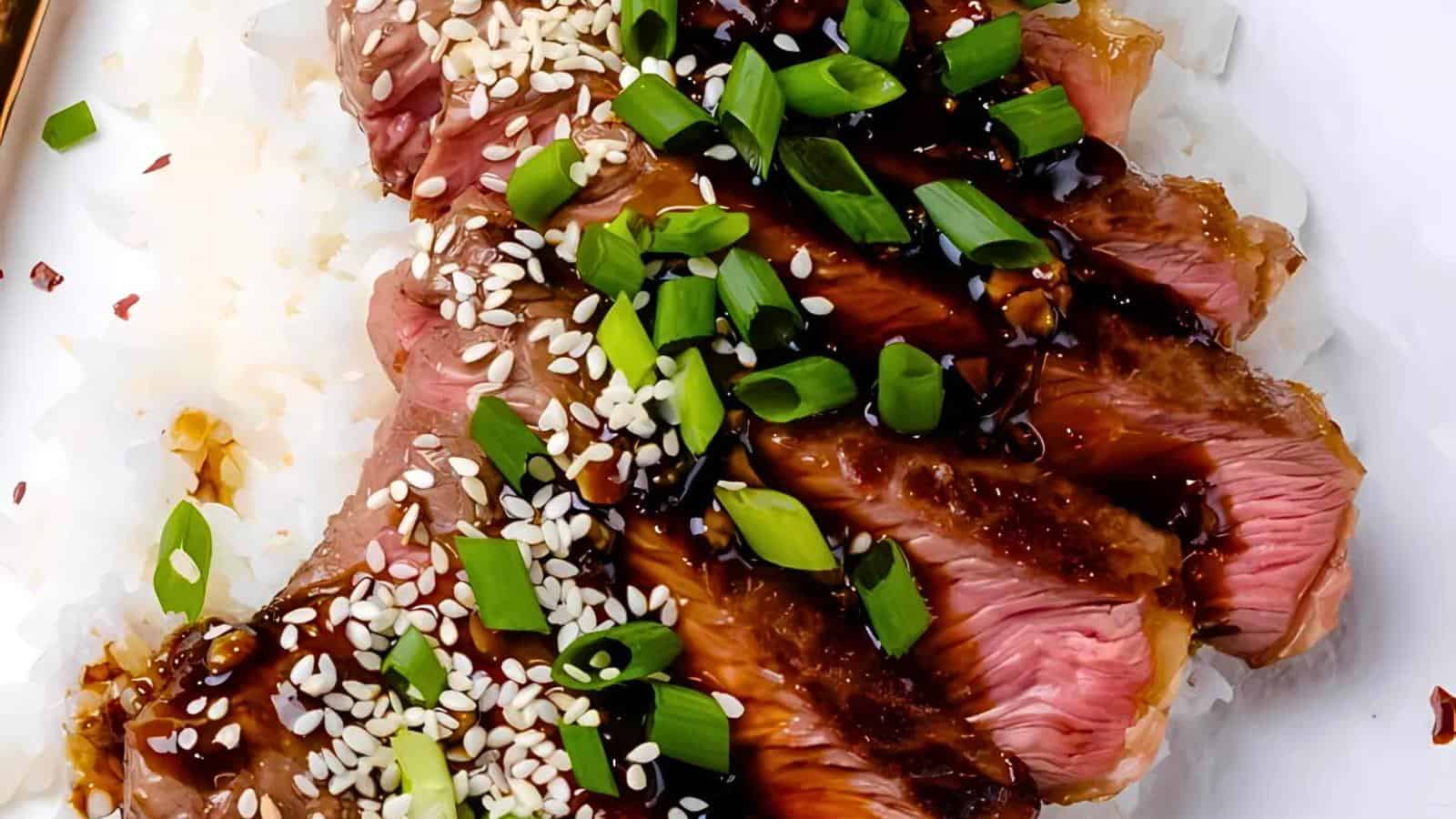 Close-up of sliced seared beef steak garnished with sesame seeds and chopped green onions, drizzled with a dark savory sauce. The steak rests on a bed of steamed white rice. The image showcases the juicy, medium-rare interior and the vibrant garnish.