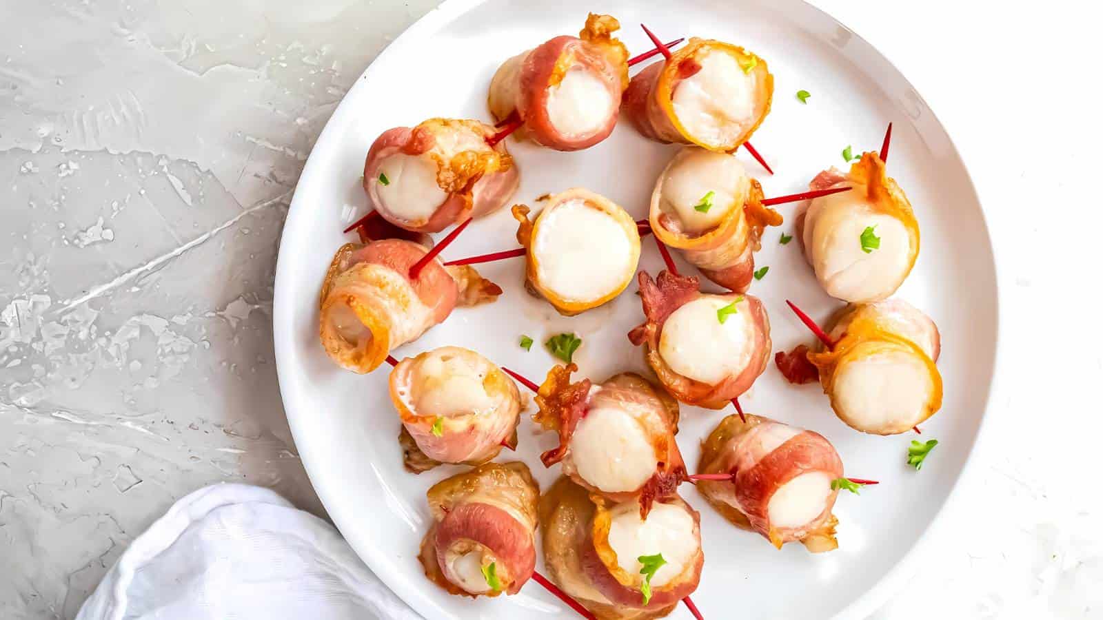 A white plate with bacon-wrapped scallops secured by red toothpicks, garnished with small pieces of green herbs.