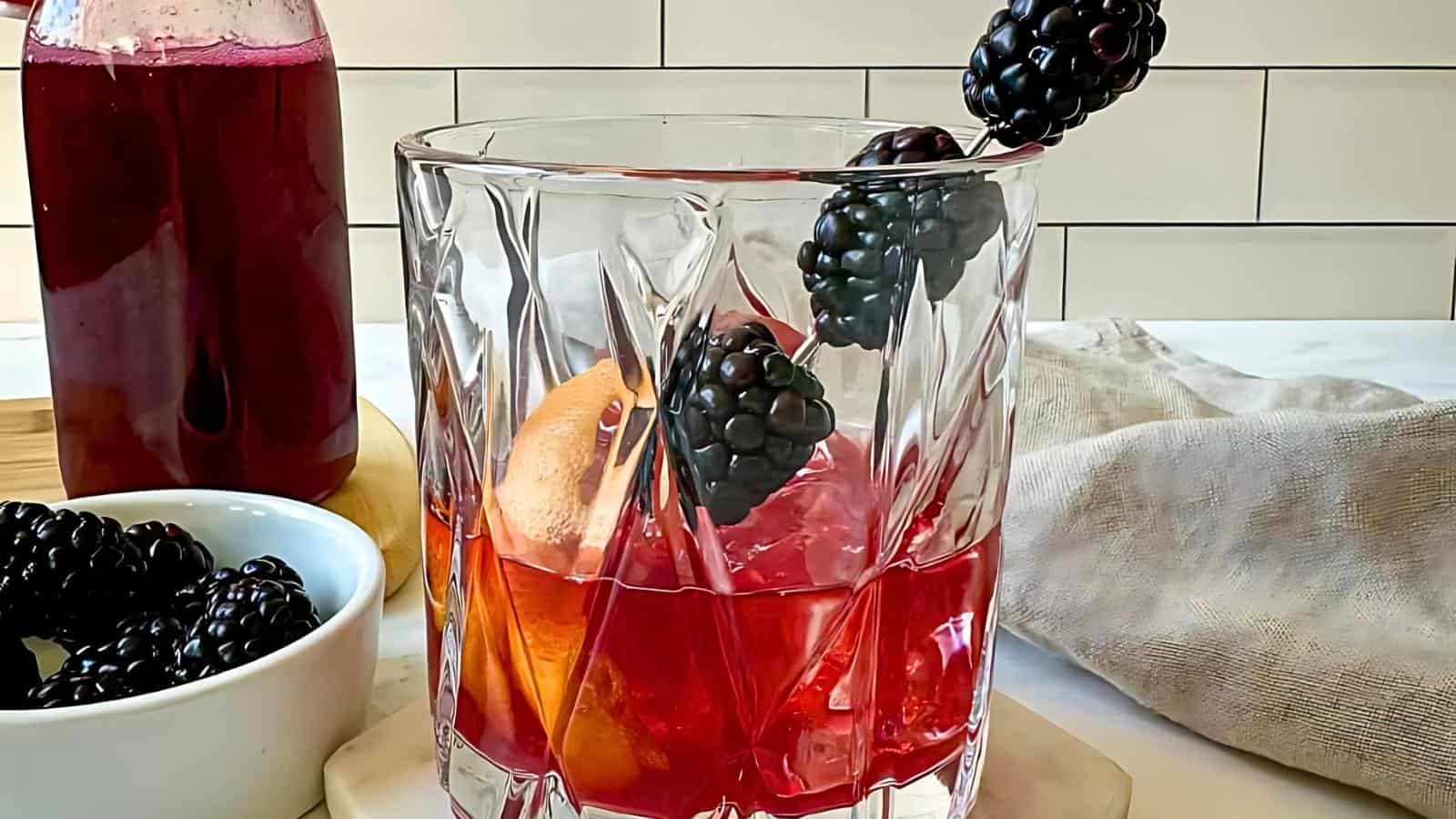 A glass filled with a red beverage, garnished with blackberries and an orange slice. Nearby are a bowl of blackberries and a bottle of the same red liquid.