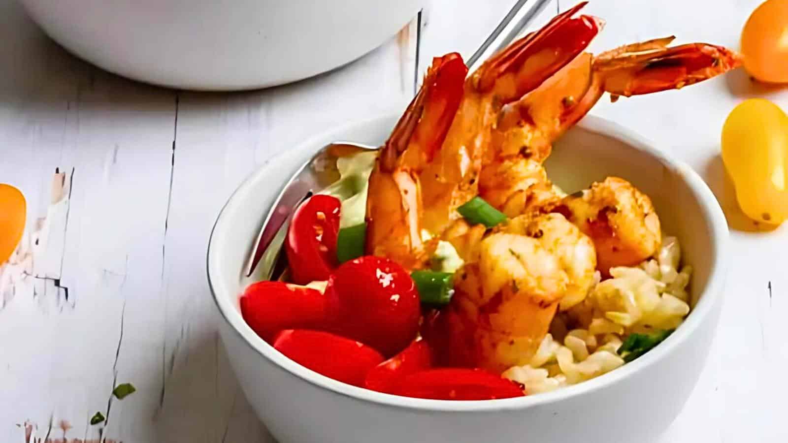 A bowl containing shrimp, red bell peppers, and rice, garnished with chopped green onions. A silver fork is placed in the bowl. Yellow cherry tomatoes are nearby on a white surface.