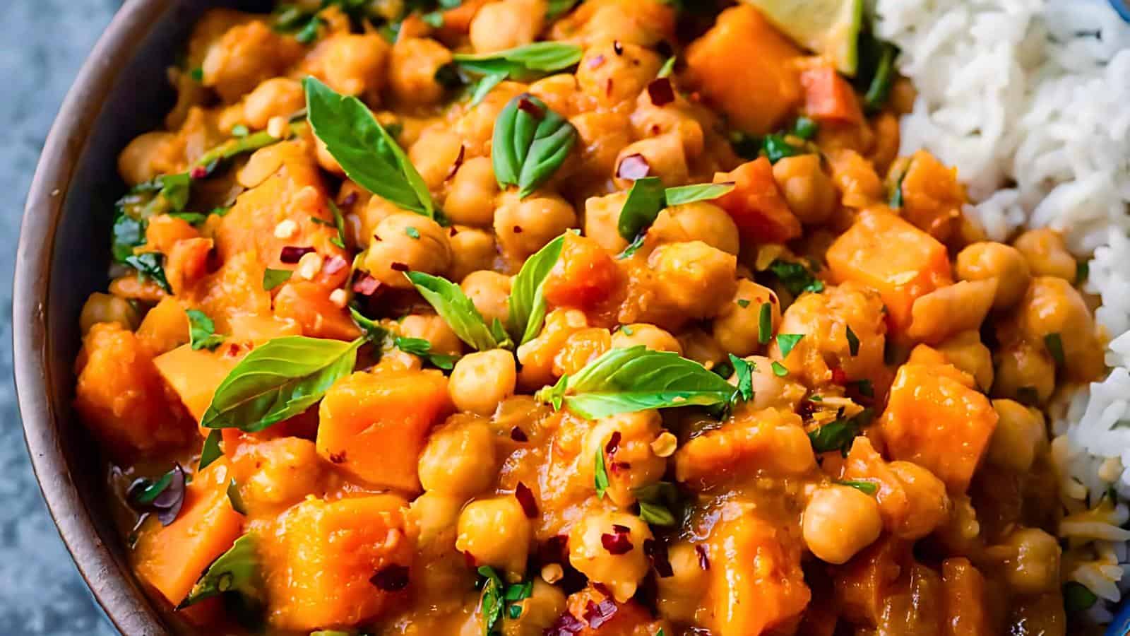 A dish with curry containing chickpeas, sweet potatoes, and fresh basil leaves, served with white rice.