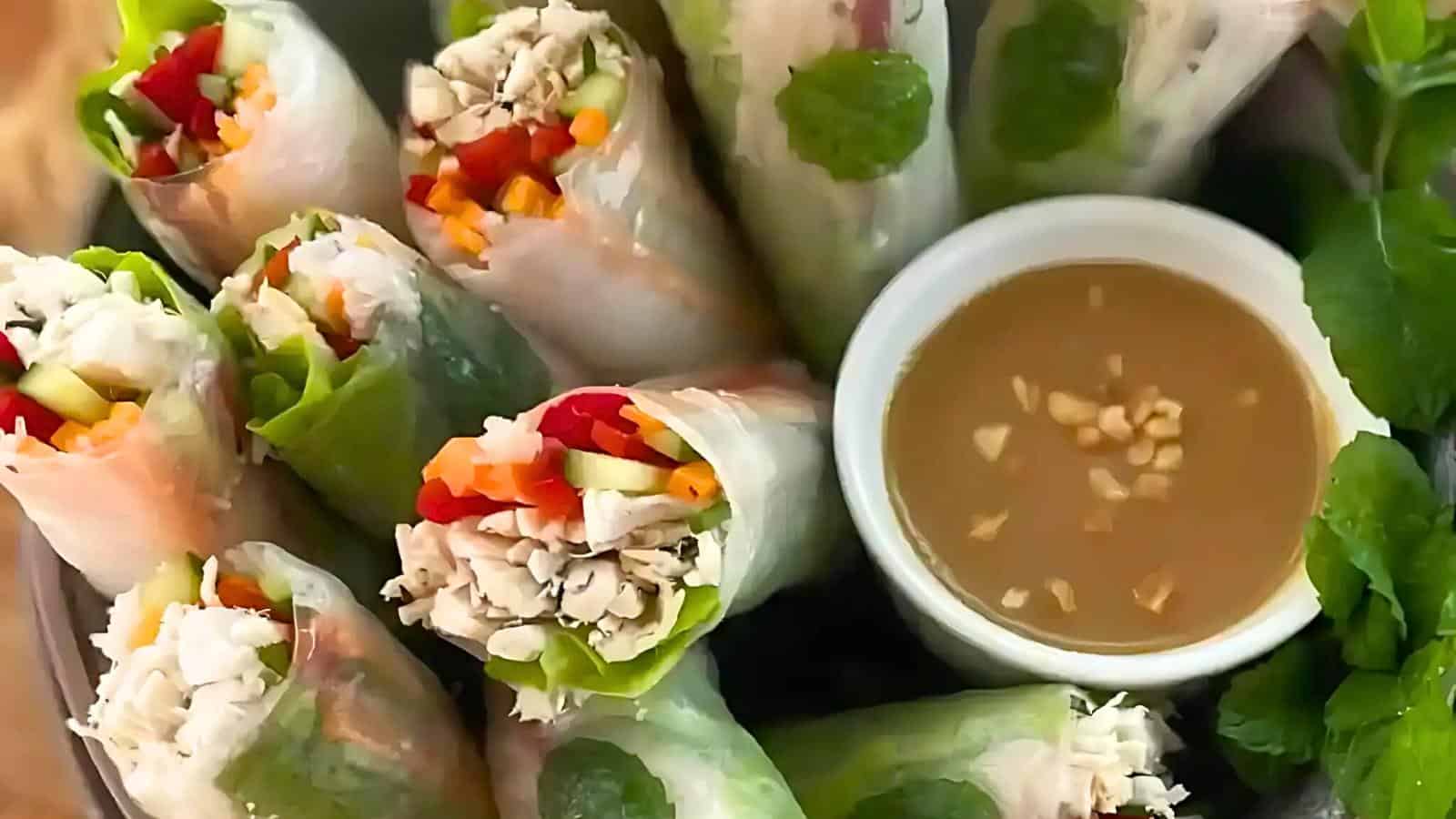 A plate of fresh spring rolls filled with vegetables and chicken, accompanied by a small bowl of dipping sauce garnished with chopped nuts.