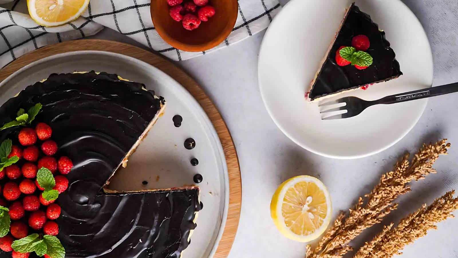 Top view of a chocolate tart with a slice on a separate plate, garnished with raspberries and mint leaves. Lemon halves, a small bowl of raspberries, and wheat stalks are also visible.