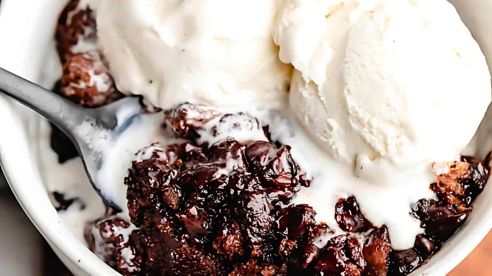 A close-up of a bowl filled with a rich, gooey chocolate dessert topped with scoops of vanilla ice cream. A spoon is seen scooping some of the chocolate mixture, blending the melted ice cream with the decadent dessert.