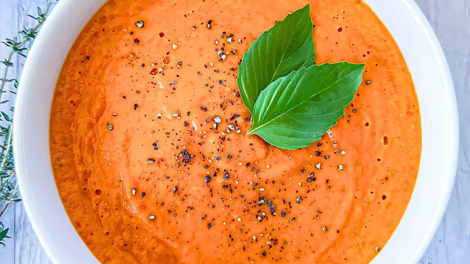 A bowl of creamy tomato soup garnished with two fresh basil leaves and a sprinkle of black pepper.