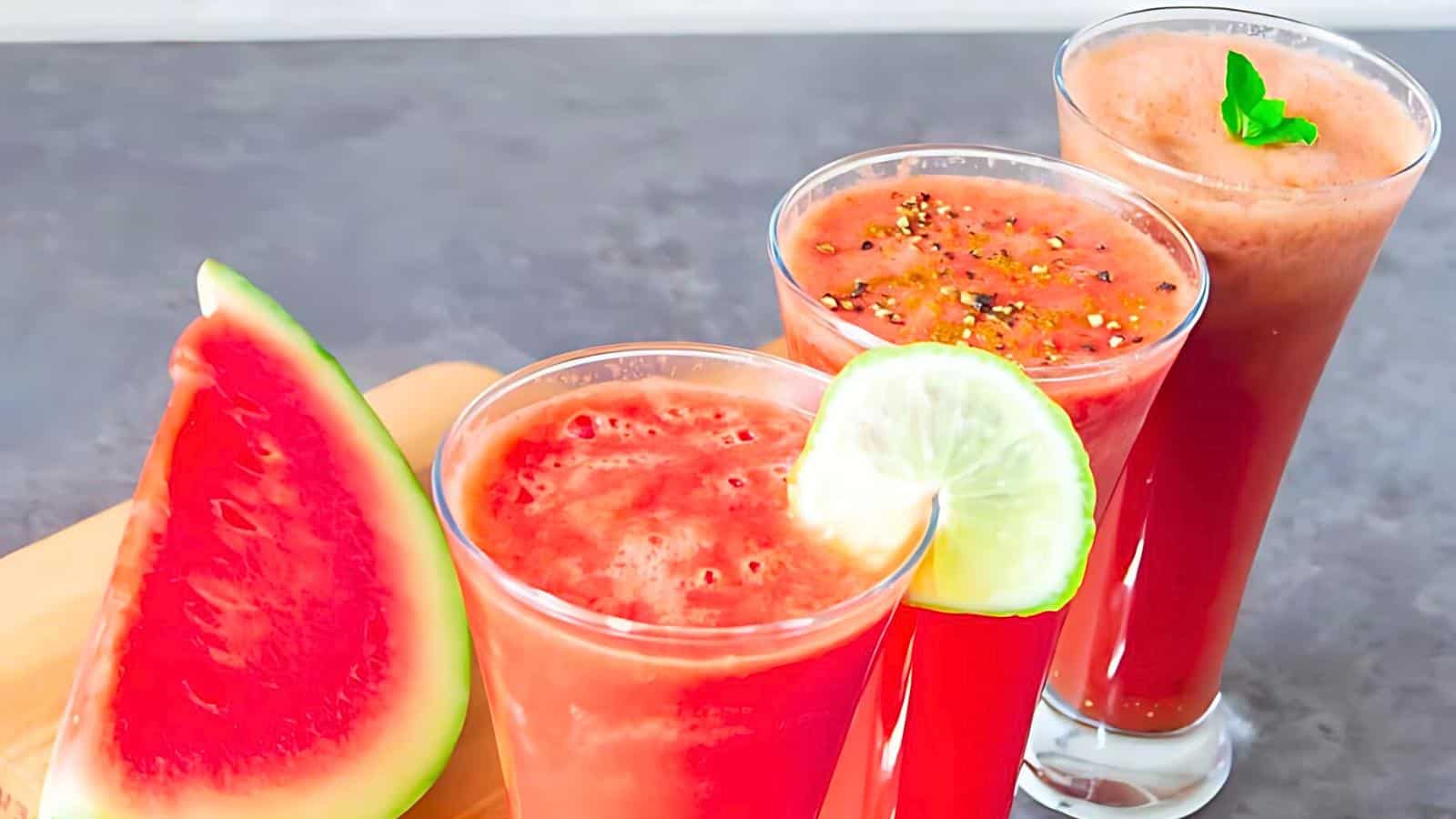 Three glasses filled with red juice, each garnished differently with lime, pepper, and mint. A slice of watermelon placed beside the glasses on a cutting board.