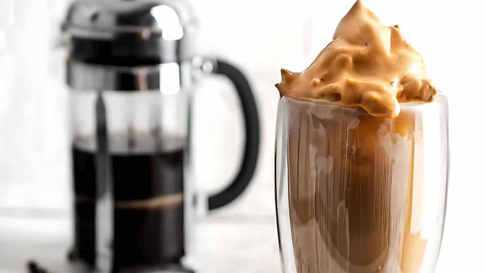 A glass cup filled with whipped coffee sits in the foreground. A French press coffee maker filled with black coffee is blurred in the background.
