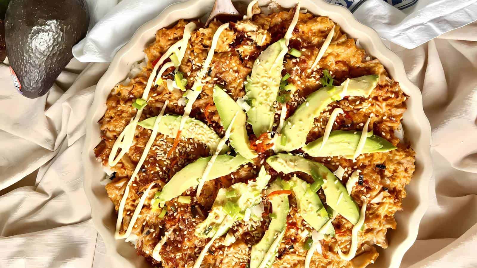 A baked dish topped with sliced avocado, drizzled with a white sauce, and sprinkled with sesame seeds.