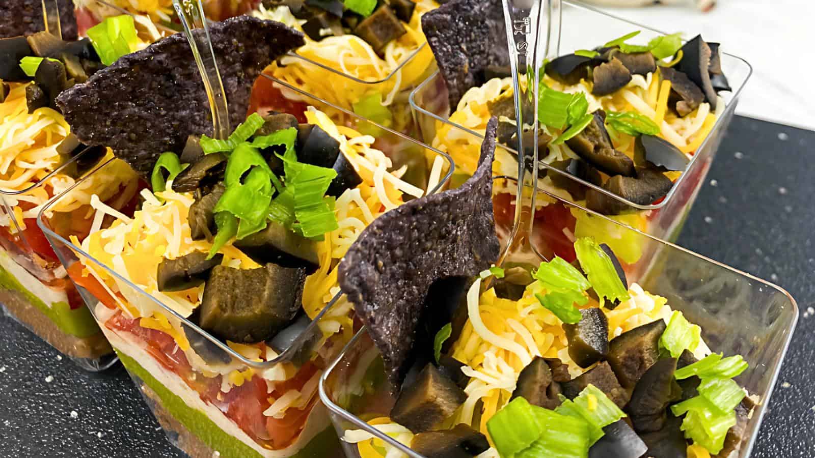 Small clear cups with layered lettuce, shredded cheese, chopped tomatoes, green onions, black olives, and a triangular blue corn tortilla chip on top.