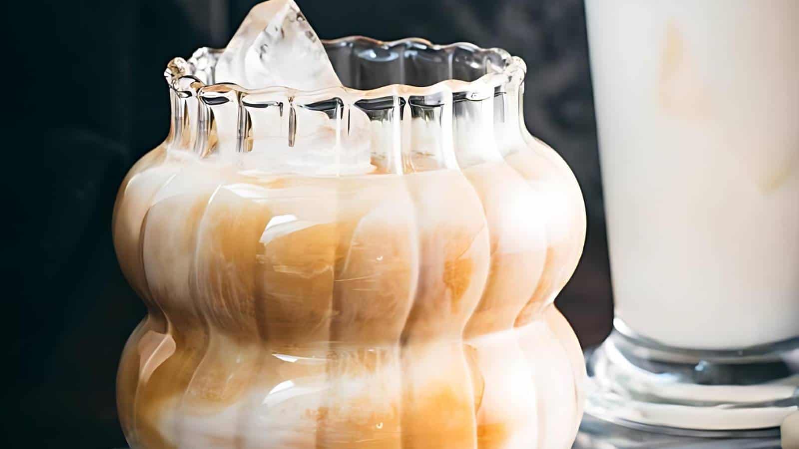 A clear, ribbed glass filled with a creamy swirled iced beverage, topped with an ice cube. Another drink is partially visible in the background.