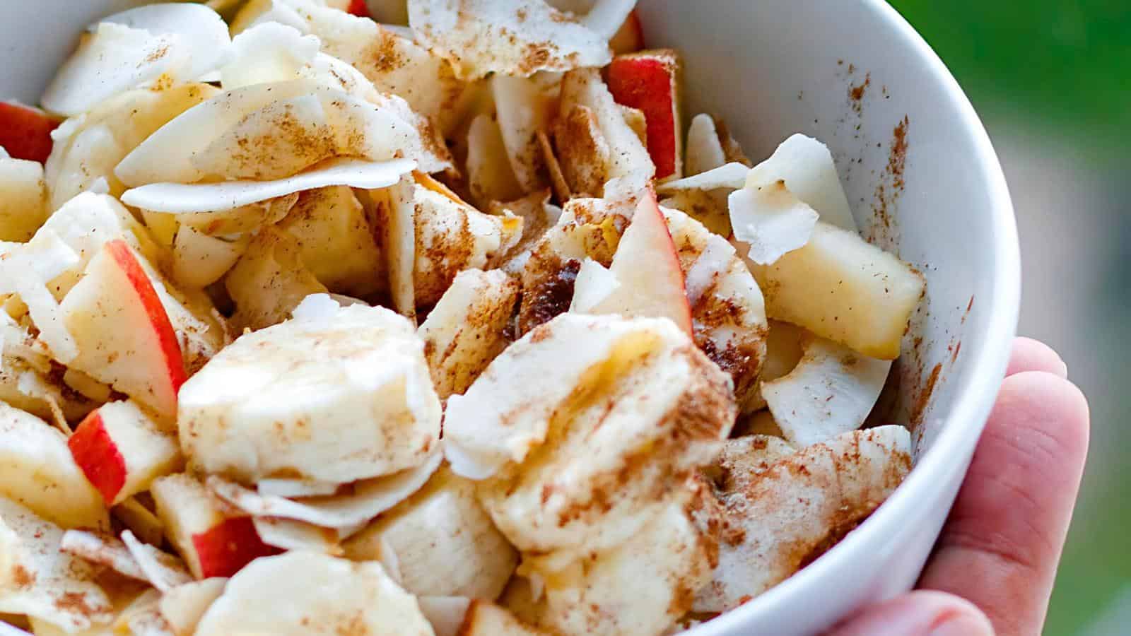 A bowl filled with sliced fruits, including bananas and apples, topped with shredded coconut and sprinkled with cinnamon. The bowl is being held outdoors, with a blurry green background.
