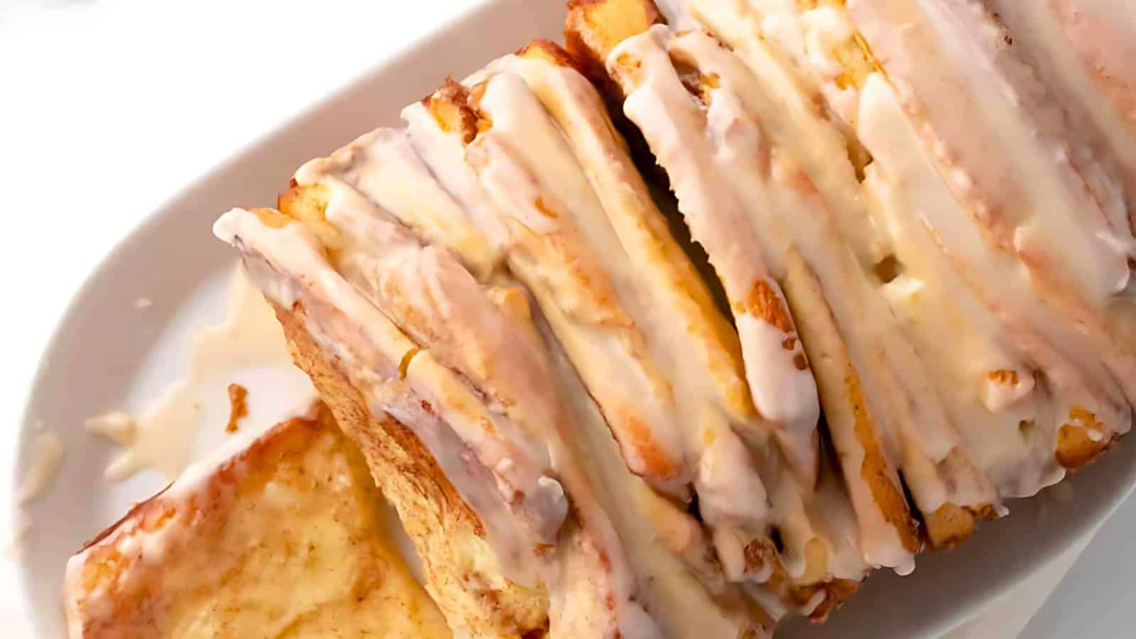 A close-up of a pull-apart bread on a white platter. The bread is layered and covered in white icing, with a golden-brown crust visible beneath the glaze.