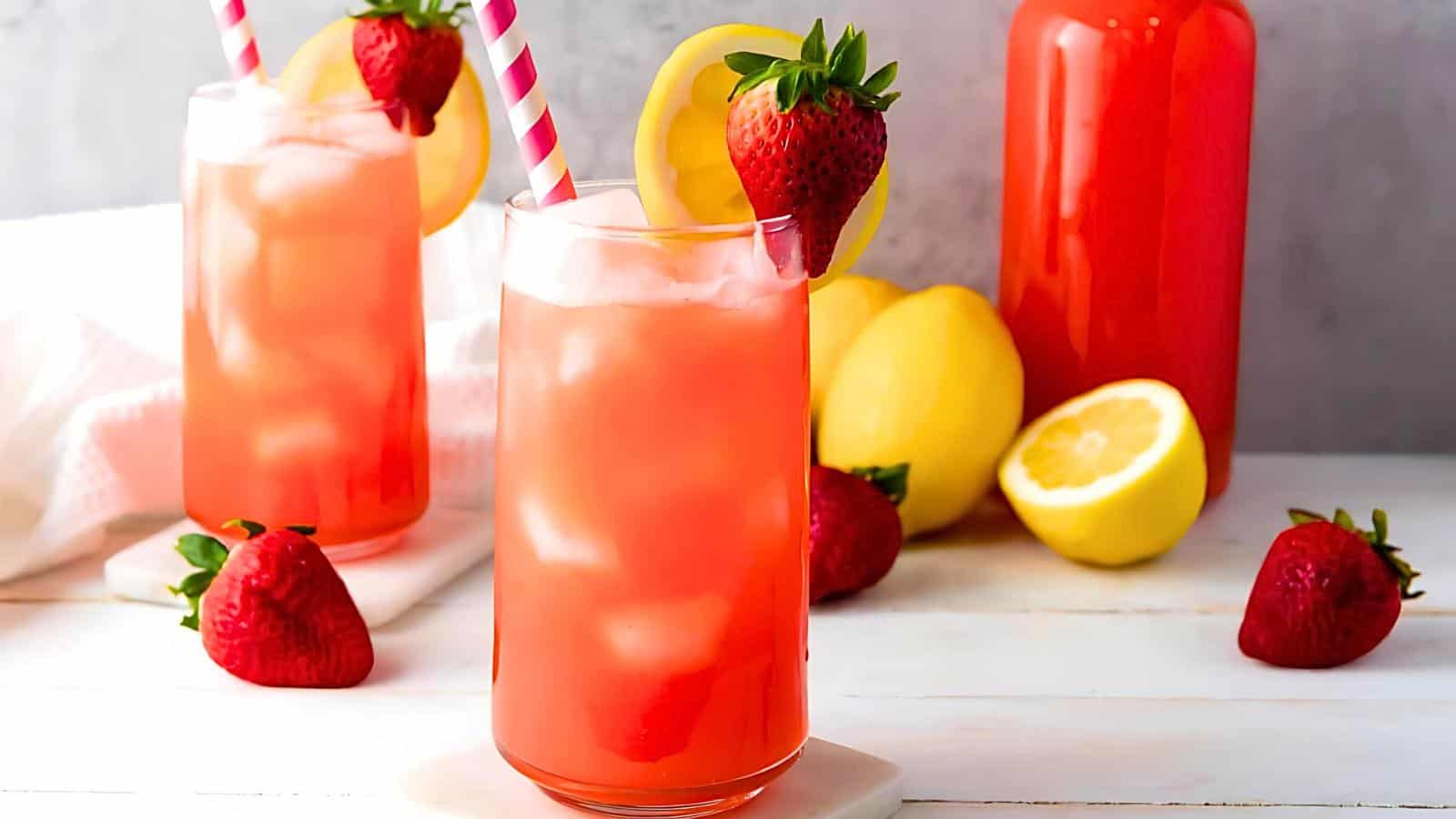 Two tall glasses of strawberry lemonade with ice, garnished with fresh strawberries and lemon slices, sit on a table. Scattered strawberries, lemons, and a bottle of the drink are in the background.