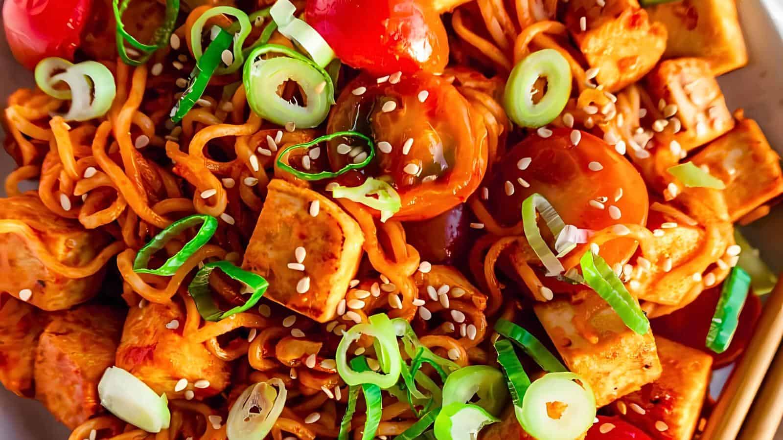 A close-up of a colorful dish featuring stir-fried noodles mixed with cubes of tofu, halved cherry tomatoes, and sliced green onions. The dish is garnished with sesame seeds and appears to be coated in a savory sauce.