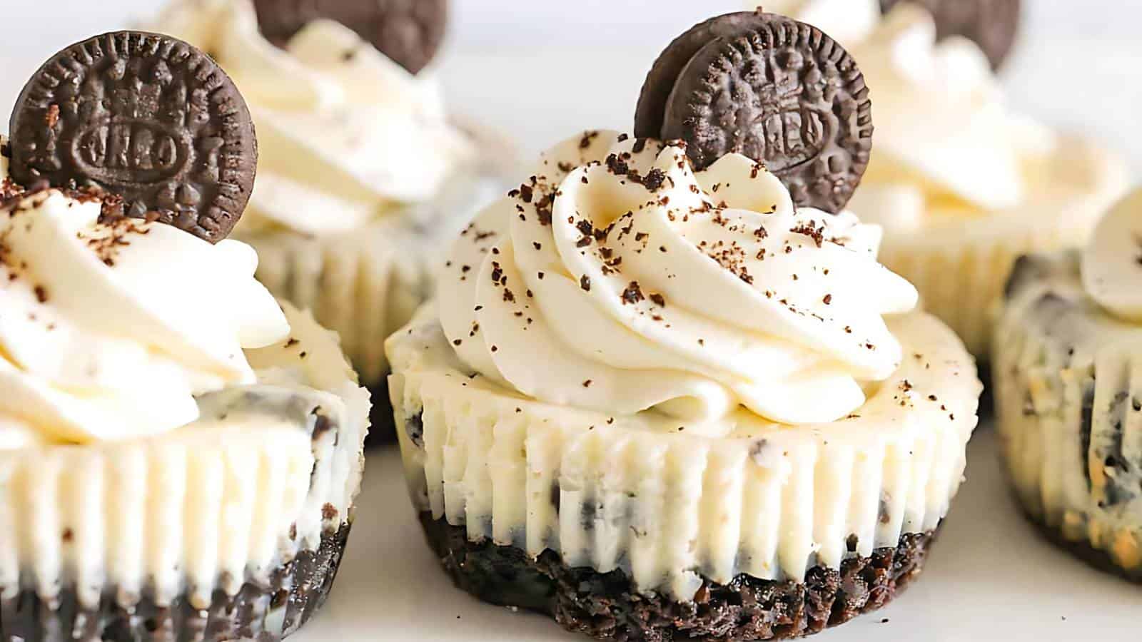 Mini Oreo cheesecakes topped with whipped cream and a mini Oreo cookie. The cheesecakes have a dark, crumbly base, creamy filling, and are decorated with chocolate crumbs. They are arranged closely together on a white surface.