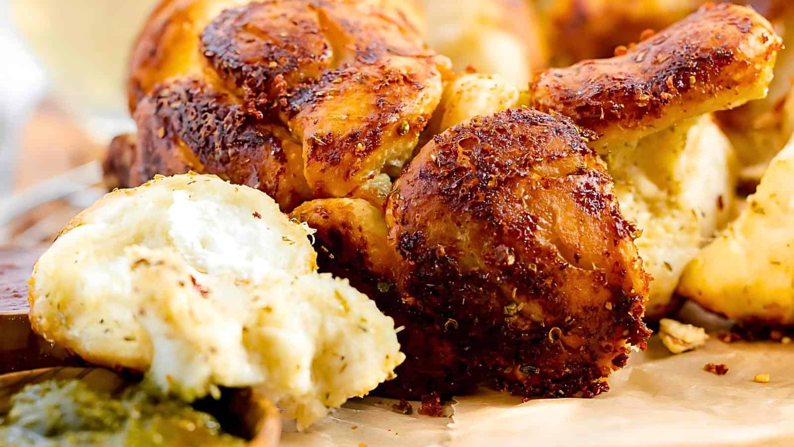 Close-up of freshly baked, golden-brown garlic bread knots sprinkled with herbs. A small dollop of green pesto is visible on the side, adding a pop of color to the warm, textured bread.