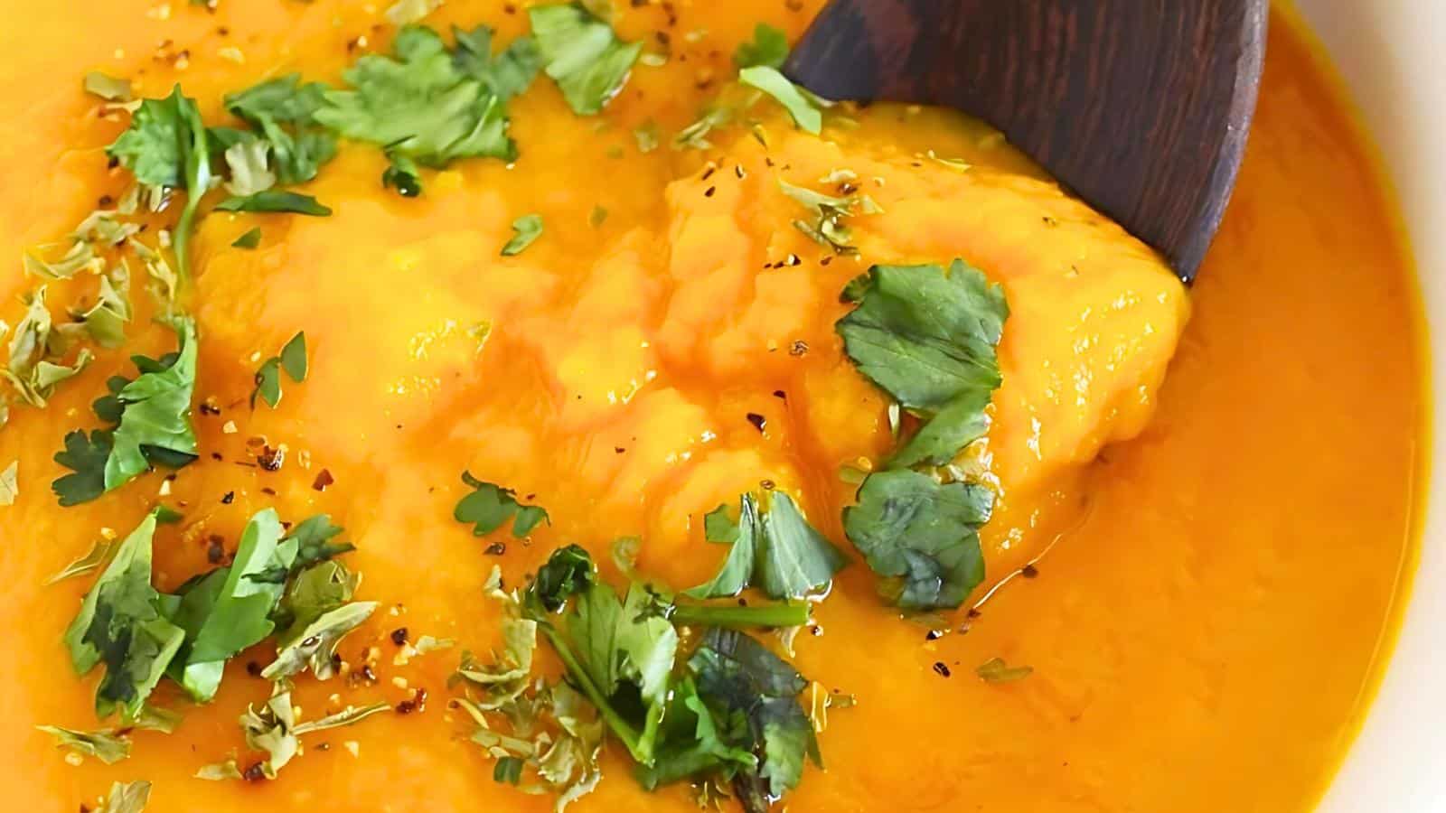 A bowl of creamy carrot soup garnished with chopped herbs and black pepper, with a wooden spoon partially immersed.