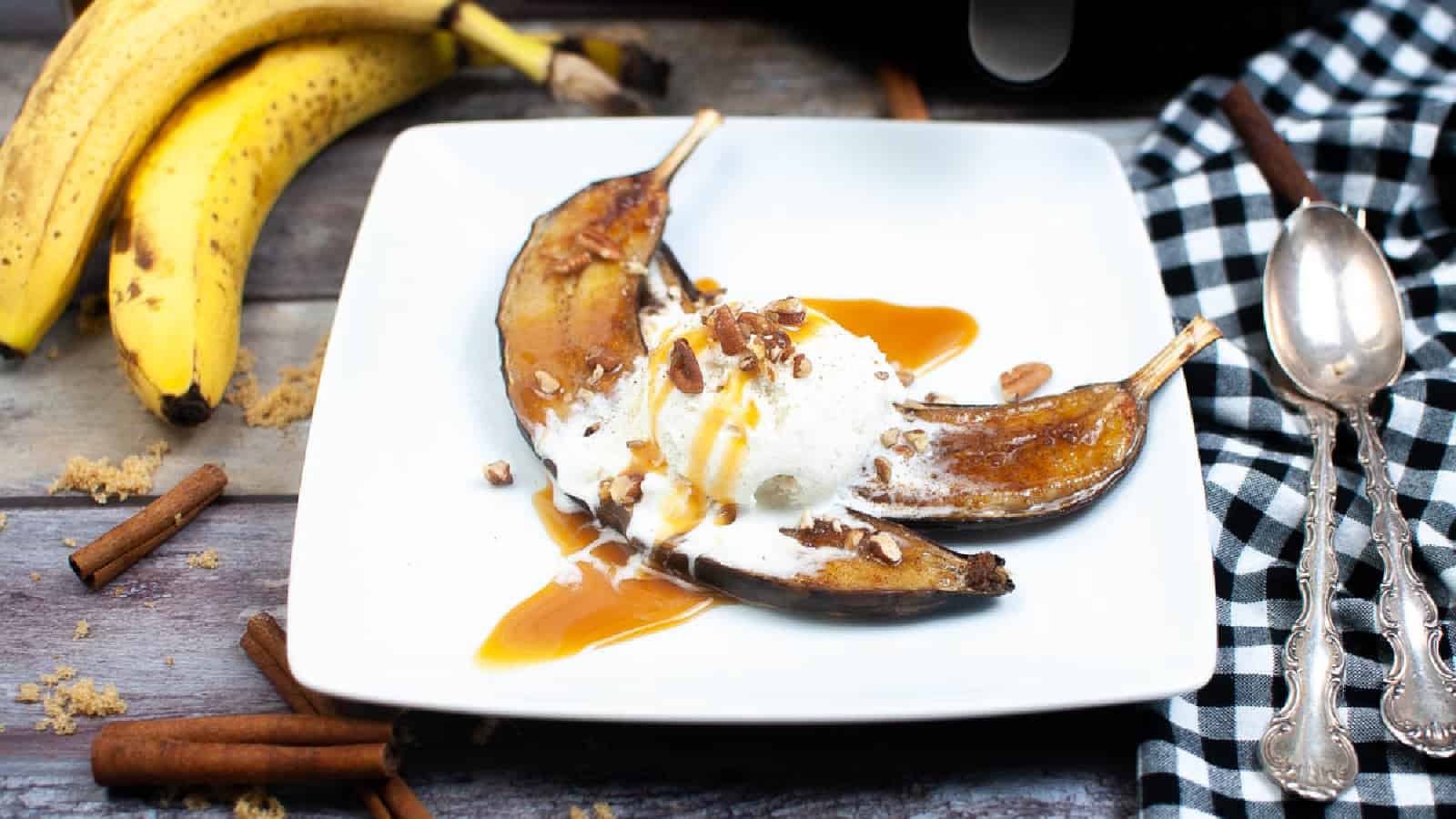 A dessert plate featuring roasted bananas drizzled with caramel sauce, topped with a scoop of vanilla ice cream and sprinkled with pecans. Fresh bananas, cinnamon sticks, a black and white checkered cloth, and silver spoons surround the plate.