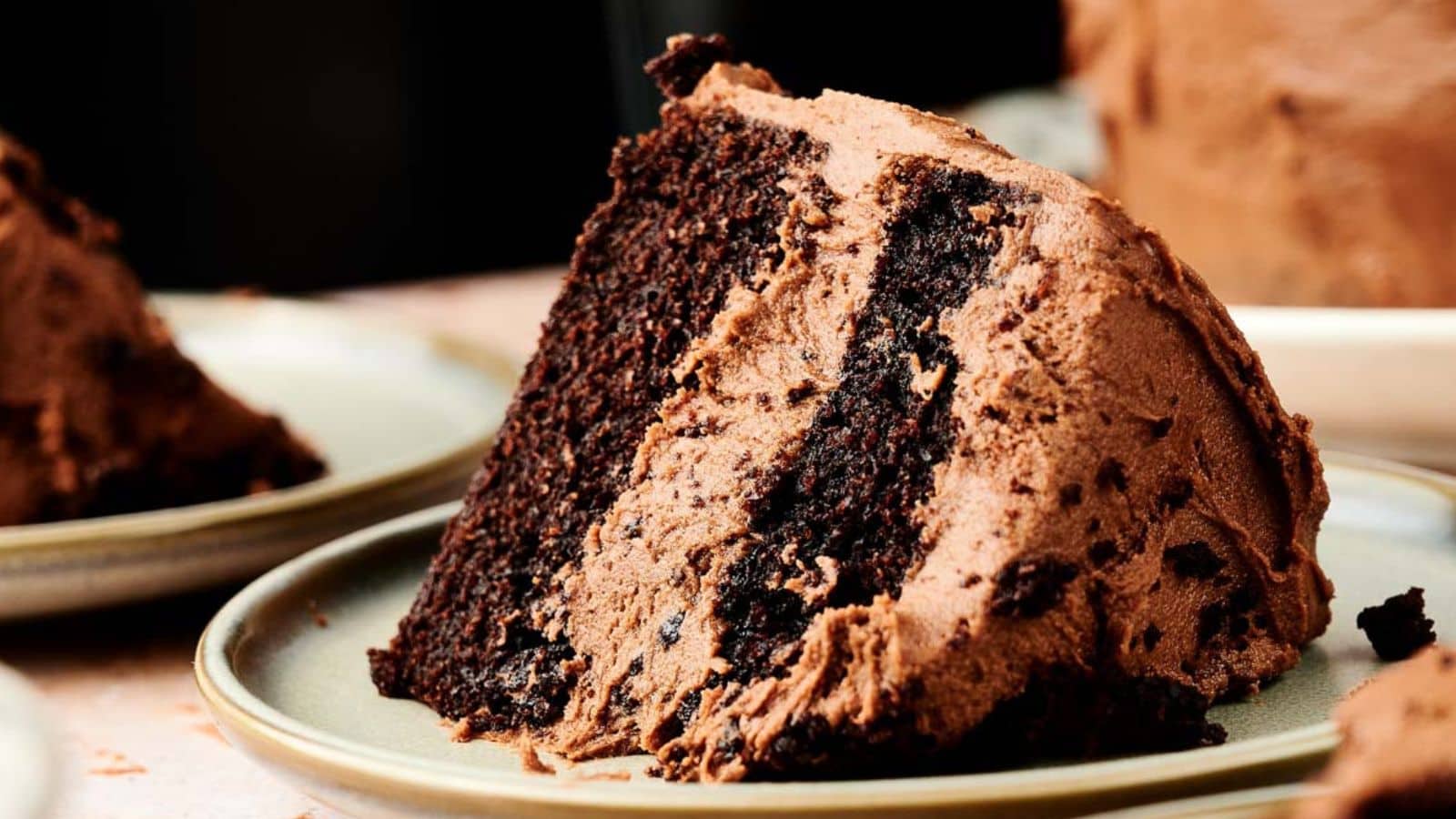 A close-up of a slice of chocolate cake with a layer of chocolate frosting and chocolate crumble, served on a light green plate.