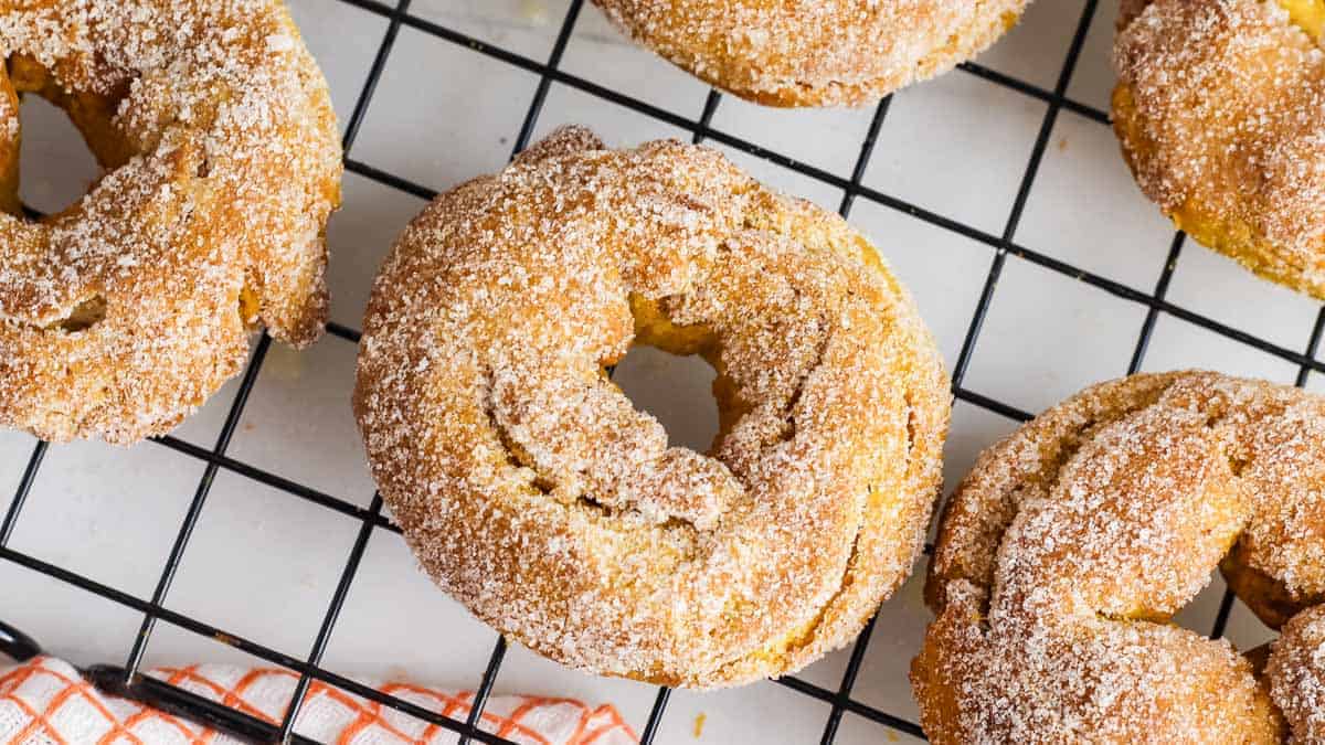 Close-up of sugar-coated donuts on a black wire cooling rack. The donuts are dusted with a generous layer of sugar and cinnamon, creating a textured, sweet appearance. An orange and white checkered cloth is partially visible at the corner.