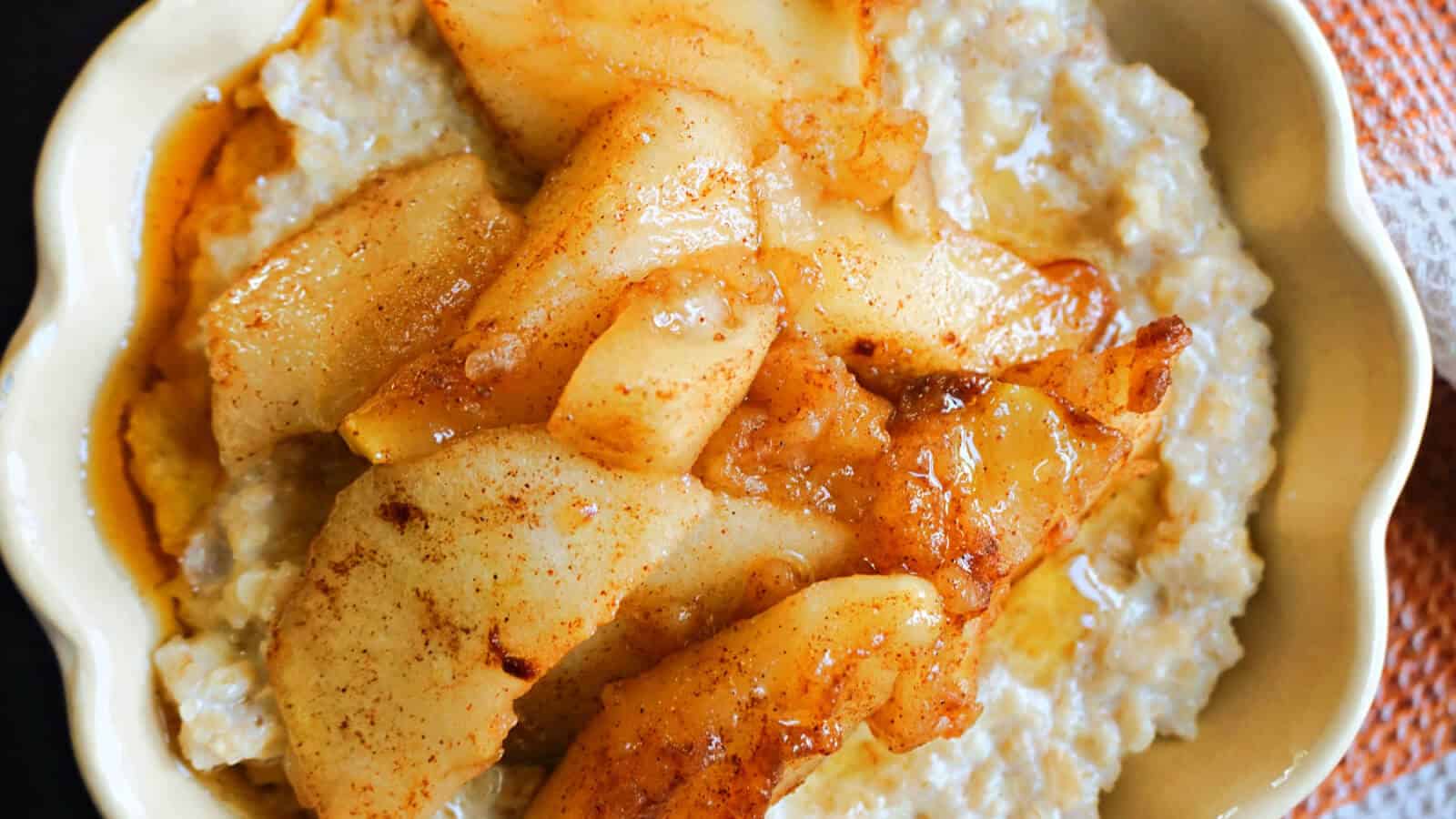 A close-up of a bowl of oatmeal topped with caramelized apple slices dusted with cinnamon. The oatmeal appears creamy, and the apples have a golden-brown hue, suggesting they were cooked with sugar and spices. The dish exudes a warm and comforting presentation.