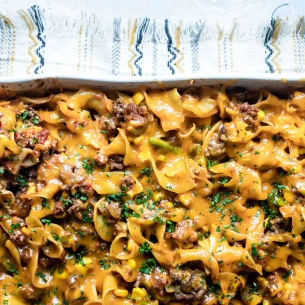 A baked casserole dish with noodles, ground meat, cheese, and garnished with chopped herbs. A patterned cloth is partially visible underneath the dish.