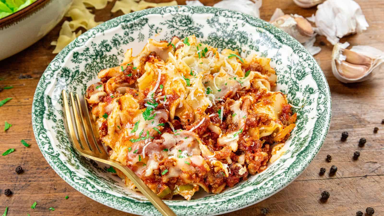 A bowl of pasta topped with meat sauce, melted cheese, and chopped herbs sits on a wooden table. A gold fork rests beside the bowl, with garlic cloves and peppercorns scattered nearby.
