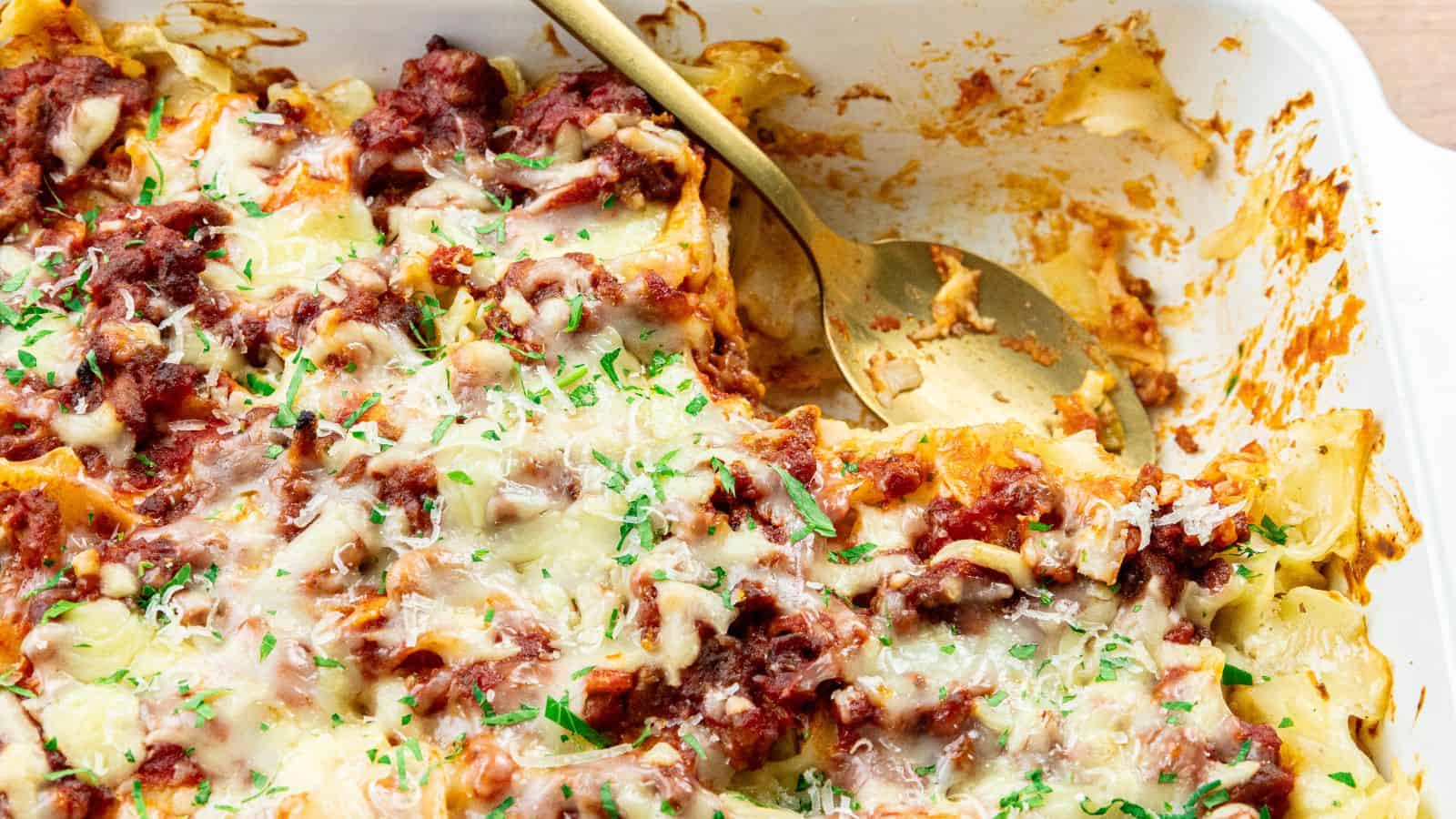 A baked pasta dish with melted cheese, tomato sauce, and herbs in a white baking dish, accompanied by a serving spoon.