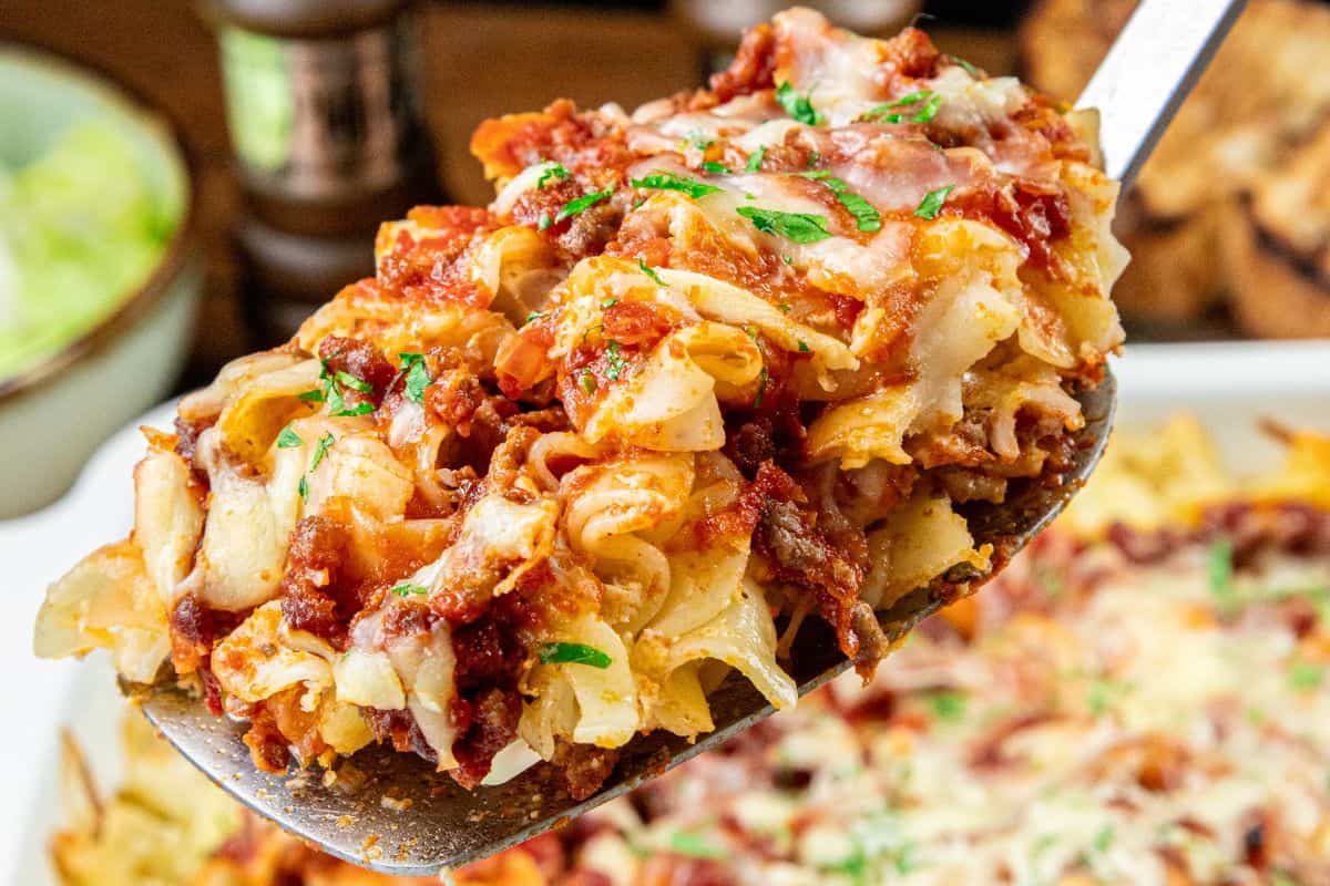 A serving spatula lifts a portion of baked pasta layered with melted cheese, tomato sauce, ground meat, and sprinkled with chopped parsley.