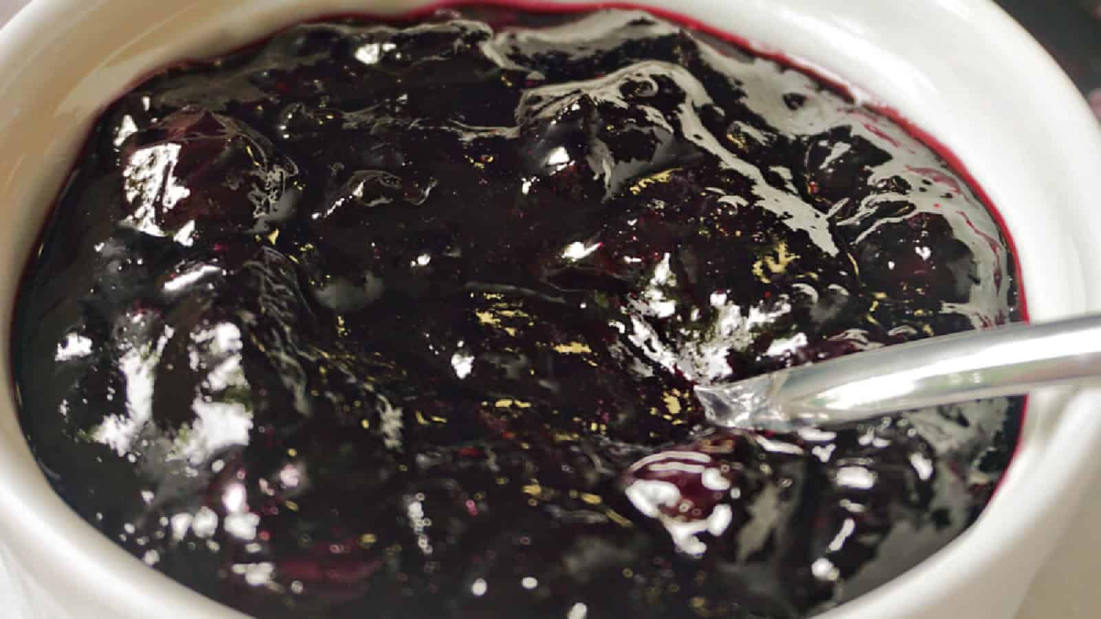 A white ramekin with blueberry jam and a teaspoon in it.